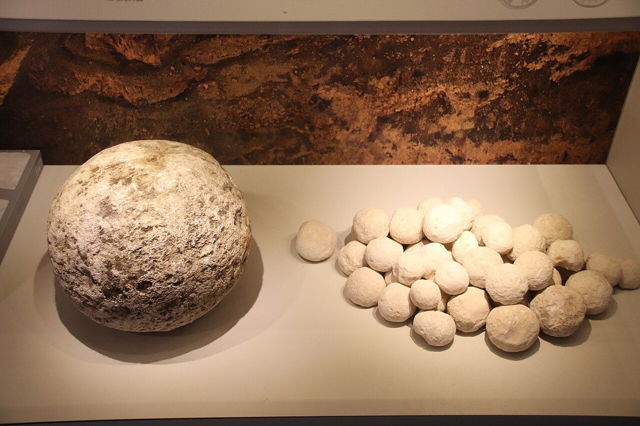 A large rounded ballista stone beside a cluster of fist-sized sling stones on a museum shelf